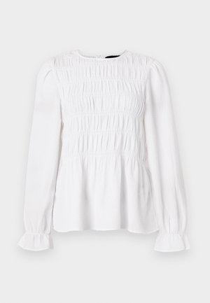 Haut à manches longues blanc avec une texture froncée, encolure ronde et poignets volants. Tissu léger avec une coupe légèrement ample.
