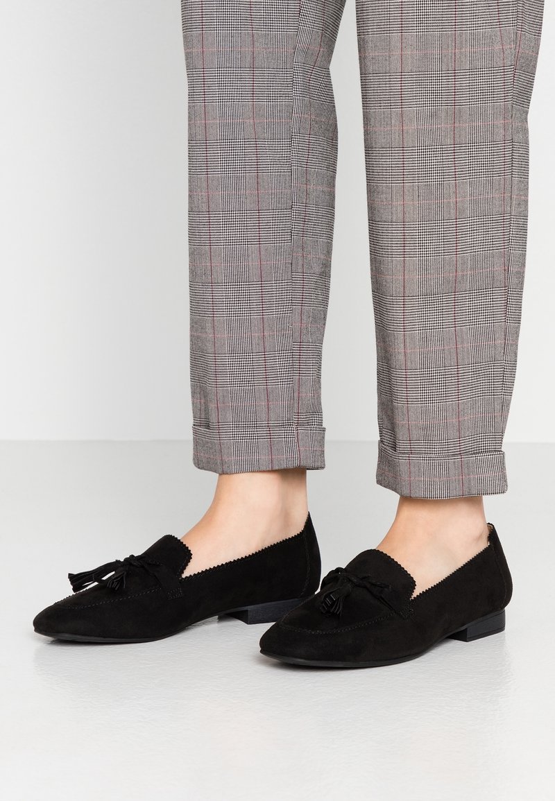 Mocassins en daim noir à talon plat, avec un nœud décoratif, des bords festonnés et une forme d'orteil en amande, assortis à un pantalon à carreaux.