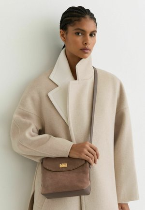 Barna szarvasbőr és bőr keresztpántos táska, füllel záródó, arany színű kiegészítőkkel, egy bézs színű, magas nyakú kabátot viselő személy által hordva.