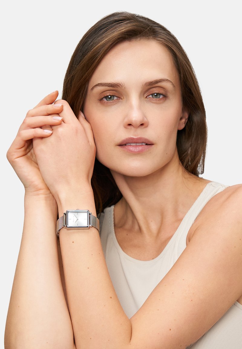 Silberne Metallmasche-Uhr mit quadratischem weißen Zifferblatt, am Handgelenk getragen, vor einem neutralen Hintergrund. Weibliches Model mit mittlerem Hautton.