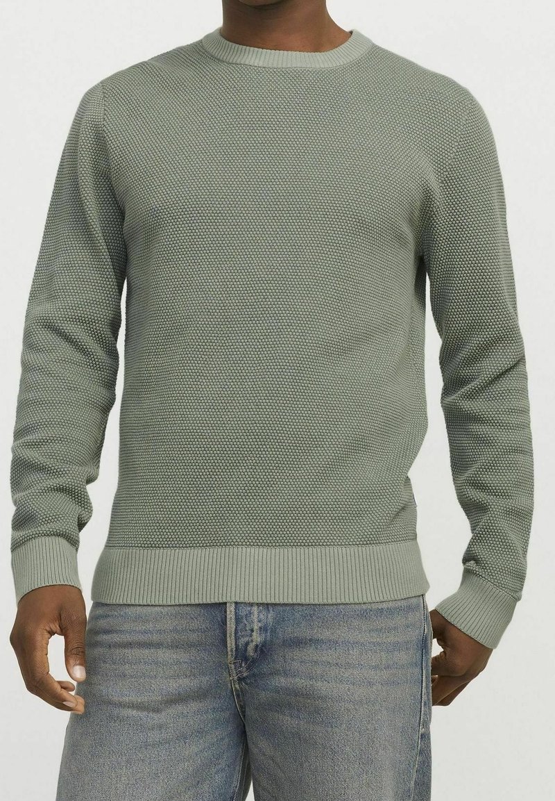 Olivgrön stickad tröja med ett strukturerat mönster, rund halsringning och ribbade ärmslut och nederkant. Bärs över blekblå raka jeans.