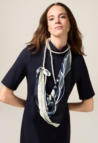 Niebiesko-kremowy jedwabny szalik z geometrycznym wzorem, zarzucony na szyję, nałożony na granatową sukienkę o wysokim dekolcie i krótkich rękawach.