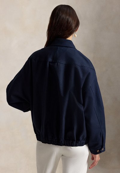 Γυναίκα με μακριά σκοτεινά μαλλιά, που φοράει ένα φαρδύ μπλε σκούρο μπουφάν με ελαστικό τελείωμα και λευκό παντελόνι, κοιτάζοντας προς τα πίσω πάνω σε απλό φόντο.