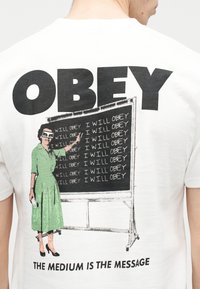 Άτομο που φοράει λευκό πουκάμισο με μεγάλο κείμενο "ΥΠΑΚΟΥΩ", εικονογράφηση γυναίκας με πράσινο φόρεμα που γράφει επανειλημμένα "ΘΑ ΥΠΑΚΟΥΩ" σε μαυροπίνακα, και φράση από κάτω.