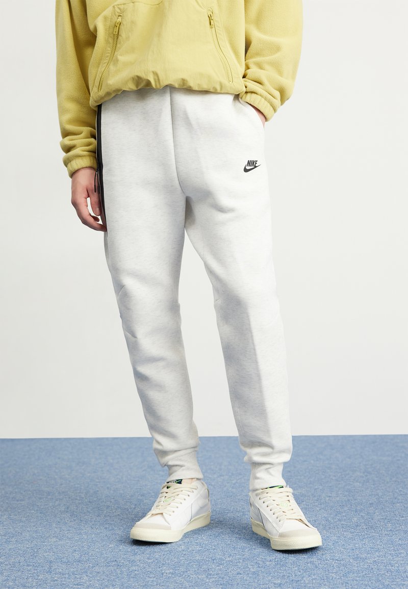 Persona que lleva pantalones de chándal Nike gris claro, zapatillas blancas y una chaqueta de forro polar amarilla con cremallera, de pie sobre una moqueta azul.