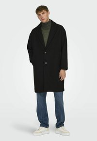Abrigo negro con solapa de muesca, cierre de botón y dos bolsillos laterales. Llevado sobre un cuello alto de punto verde y combinado con jeans azules y zapatillas blancas.