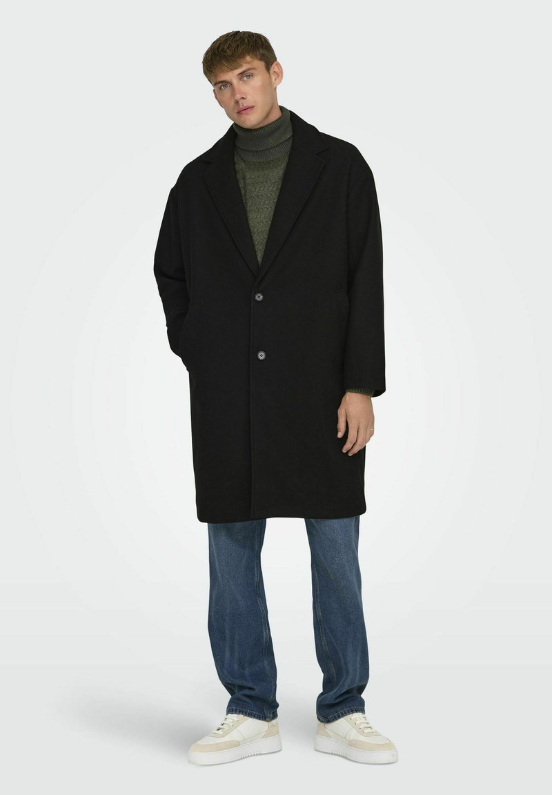 Abrigo negro con solapa de muesca, cierre de botón y dos bolsillos laterales. Llevado sobre un cuello alto de punto verde y combinado con jeans azules y zapatillas blancas.