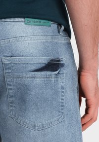 Jeans de mezclilla azul claro con un corte recto, que presentan un bolsillo trasero, una etiqueta de logo en teal y un ligero desgaste cerca del área del bolsillo.