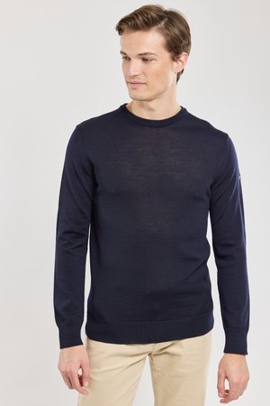 Giovane uomo che guarda verso destra, indossa un maglione blu navy a maniche lunghe e pantaloni beige su uno sfondo chiaro e semplice.