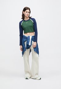 Zöld, rövidített, hosszú ujjú felső, sötétkék ujjakban, fehér díszítéssel. A modellen világos bézs széles lábú nadrág és a derekára kötött kék dzseki van.