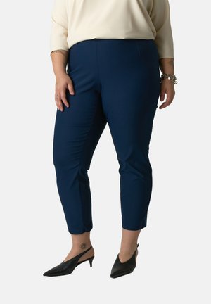 Pantalons cropped bleu marine avec une texture lisse, des détails sur les panneaux latéraux et une fermeture éclair sur le côté, accompagnés de talons aiguilles noirs.