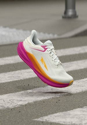 Weißer Laufschuh mit pinker und oranger Sohle, schwebt über einem Zebrastreifen auf Asphalt.