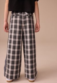 Pantalon large à carreaux noirs et blancs avec une texture légère, doté de bandes latérales et de deux poches arrière. Porté avec des baskets blanches.