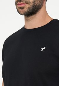 Άνδρας με μούσι φοράει μαύρο T-shirt με στρογγυλή λαιμόκοψη και μικρό λευκό λογότυπο πουλιού στο αριστερό μέρος του στήθους.
