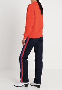 Sudadera roja con puños acanalados, combinada con jeans de mezclilla oscuros que tienen rayas verticales rojas y azules y botas de tobillo blancas con tacón cuadrado.