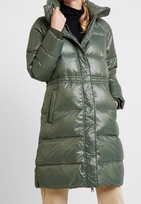 Mujer vestida con un abrigo de invierno acolchado largo en color oliva con un cuello alto, cierre con cremallera y botones a presión, y bolsillos laterales, sujetando el cuello con una mano.