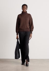 Maglione marrone lavorato a maglia con collo alto e maniche ampie abbinato a pantaloni neri con righe sottili. La modella tiene una borsa a tracolla nera e indossa scarpe basse.