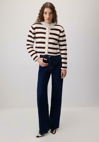 O tânără cu păr creț, purtând un pulover butonat cu dungi maro și albe, blugi evazați de culoare bleumarin și pantofi cu imprimeu de vacă.