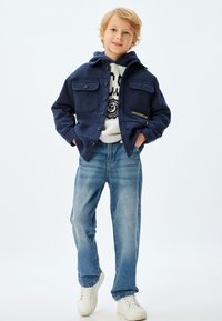 Marinejakke med to brystlommer, knaplukning og en hættetrøje nedenunder. Lys denimbukser og hvide sneakers. Teksten "UPPER MIDWEST" på jakken.