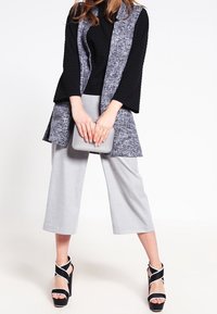 Vrouw in grijze wijde, cropped broek, zwarte top met uitlopende mouwen, gestructureerde grijze vest, zwart-witte plateauhakken, met een grijze clutch in de hand.