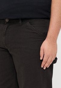 Right hand resting on thigh, wearing black pants and a black shirt, with visible pocket and belt loop details.