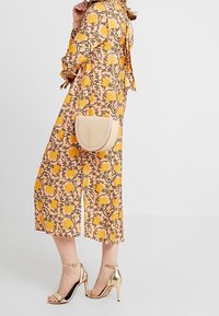 Kobieta w żółtej kwiecistej sukience midi i złotych sandałach na wysokim obcasie, trzymająca beżową torebkę w kształcie półksiężyca.