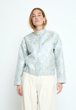 Naine, kes kannab helesinist ja kuldset mustrilist kõrge kaelusega pikka varrukatega jopet ning kreemjasid laiade säärega pükse, poseerib valge tausta ees.