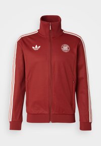 Crvena jakna na zip od mekog materijala, s bijelim Adidas trakama na rukavima, logotipom na prsima te rebrastim manžetama i donjim dijelom.