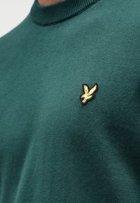 Πράσινο πλεκτό πουλόβερ με στρογγυλή λαιμόκοψη, με μικρό χρυσό λογότυπο πουλιού κεντημένο στο αριστερό στήθος. Λείο ύφασμα.