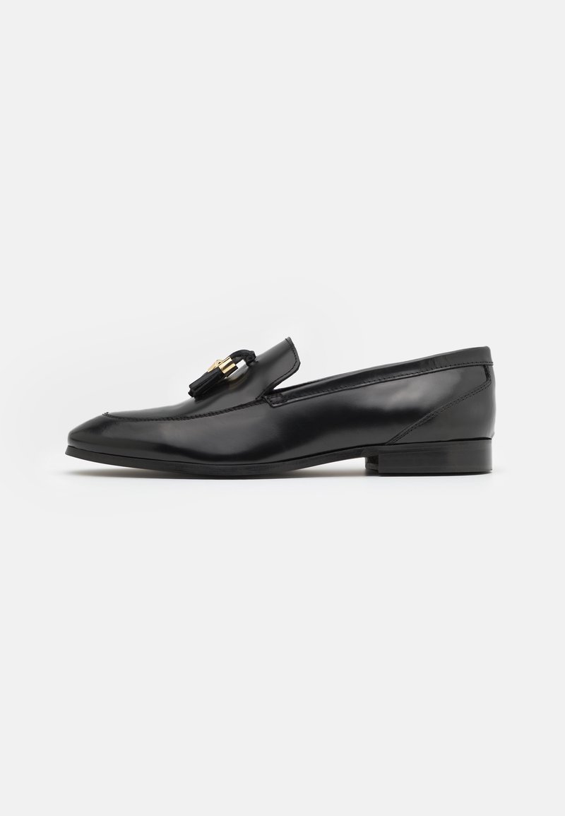 Mocassin en cuir noir brillant avec petit talon et pompon noir avec des accents dorés sur le dessus, vu de côté sur fond blanc.