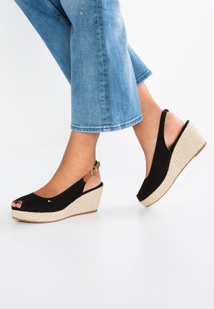 Jambes portant un jean bleu et des sandales compensées noires à bout ouvert avec une plateforme beige tissée et des brides à boucle à la cheville.