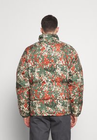 Homme portant une doudoune à motif camouflage multicolore vert, orange et beige, vu de dos sur un fond uni.