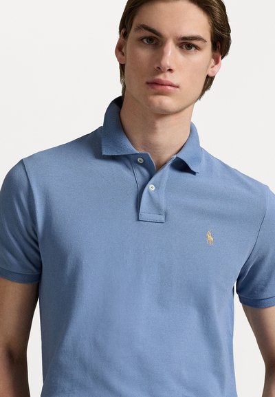 Polo de color azul claro hecho de tela texturizada. Cuenta con cuello tipo polo, tapeta de dos botones y un pequeño logo bordado en dorado.