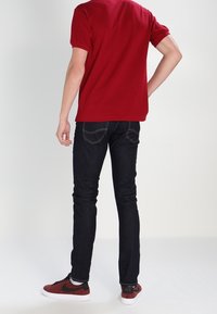 Chemise rouge à manches courtes, jean slim bleu foncé, et baskets rouges avec des accents noirs. Coutures visibles sur le jean et logo sur la chaussure.