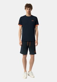 T-shirt blu navy con dettagli arancioni e logo, abbinato a pantaloni corti blu navy con cordino arancione e dettagli del logo, completato con sneakers bianche.