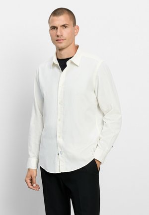 Mann mit kurzen Haaren, der ein weißes Hemd mit Knöpfen über einem schwarzen Shirt trägt, steht mit einer Hand in der Tasche vor einem einfarbigen Hintergrund.