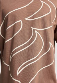 T-shirt marrone caratterizzato da un tessuto testurizzato con un motivo astratto color crema a spirale. Il design presenta linee morbide e una vestibilità rilassata.