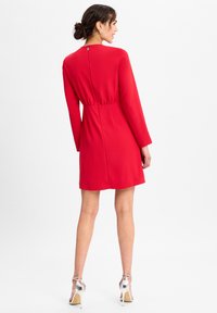 Vrouw die zijwaarts staat, gekleed in een rood jurkje met lange mouwen en zilveren sandalen met hoge hakken, tegen een effen witte achtergrond.