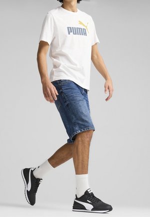Camiseta blanca con el logotipo azul de "PUMA" y un emblema amarillo de gato, combinada con pantalones cortos de mezclilla azul hasta la rodilla y zapatillas deportivas negras con acentos blancos.