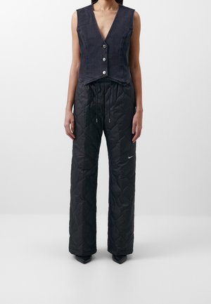 Kobieta w bezrękawniku z czarnego denimu z srebrnymi guzikami oraz w czarnych pikowanych szerokich spodniach z białym logo Nike na lewej nodze.