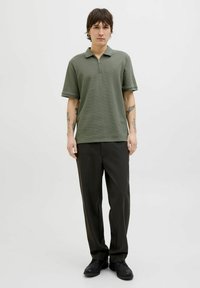 Polo shirt verde oliva con colletto a mezza zip, maniche corte e finitura testurizzata, abbinato a pantaloni scuri e calzature nere.