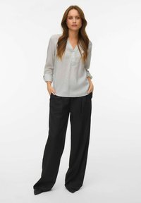 Femme aux longs cheveux portant une blouse légère à rayures avec les manches retroussées et un pantalon large noir ample, debout les mains dans les poches.