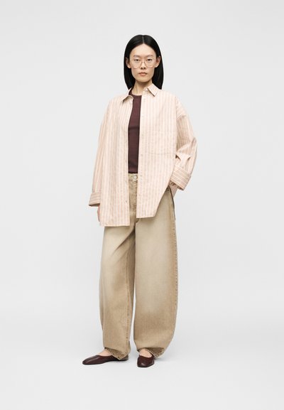 Personne aux cheveux noirs raides et portant des lunettes, vêtue d'une chemise beige rayée ample, d'un haut marron, d'un pantalon large beige et de chaussures marron foncé, debout immobile.