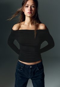 Zwarte off-shoulder top met lange mouwen, gecombineerd met donkerblauwe denim jeans met een subtiele luipaardprint en decoratieve accenten op de zakken.