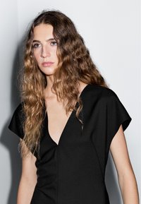 Svart V-ringad klänning med korta ärmar, tillverkad i ett slätt tyg, med en figurnära siluett och subtila sömmar. Neutral bakgrund.