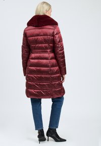 Vrouw met haar rug naar ons toe, gekleed in een lange, glanzende bordeauxrode pufferjas met een bontkraag, blauwe jeans en zwarte enkellaarsjes met een hak.