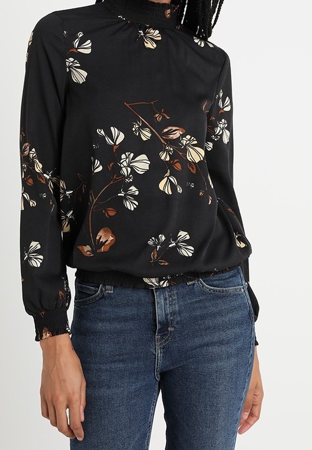 Blouse noire à manches longues avec imprimé floral beige et marron, col et poignets froncés, portée avec un jean en denim bleu.