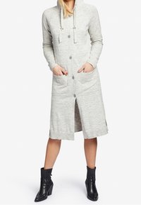 Femme portant une robe cardigan à capuche gris clair à boutons avec poches avant et des bottines noires, se tenant les mains dans les poches.