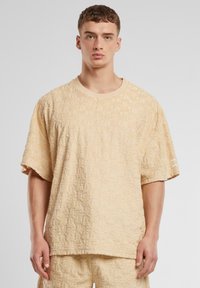 Beige strukturované tričko s krátkým rukávem s embosovaným vzorem, kulatým výstřihem a malým logem na rukávu. Zaoblený design spodního lemu.