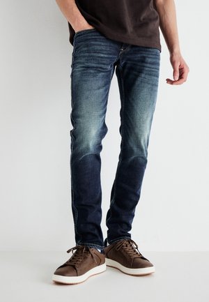 Man wearing faded blue slim-fit jeans and brown sneakers, standing with one hand in pocket against a plain background.